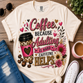A cream-colored graphic t-shirt featuring a 'Coffee Because Adulting Is Hard Caffeine Helps' design with a leopard-print coffee cup, hearts, and flowers, styled in a flat-lay with jeans, sunglasses, a cup of coffee, and sneakers on a wooden background.