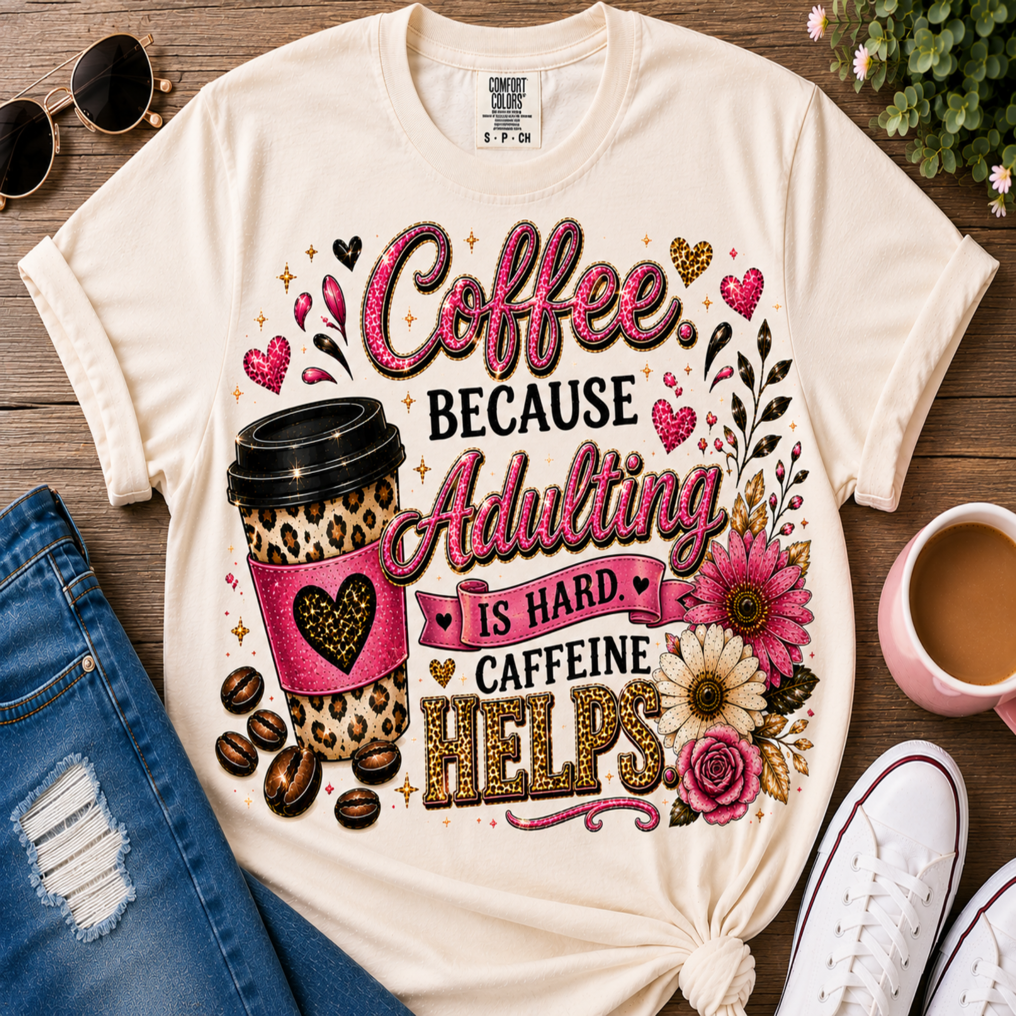 A cream-colored graphic t-shirt featuring a 'Coffee Because Adulting Is Hard Caffeine Helps' design with a leopard-print coffee cup, hearts, and flowers, styled in a flat-lay with jeans, sunglasses, a cup of coffee, and sneakers on a wooden background.
