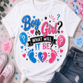 A white graphic T-shirt laid flat on a light wood surface with pink and blue baby-themed props. The shirt reads “Boy or Girl? WHAT WILL IT BE?” in blue, pink, and black lettering, with balloons, hearts, stars, question marks, and baby footprints.