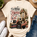 A cream-colored graphic t-shirt featuring a cat-themed design with the text 'A HOUSE IS NOT A Home WITHOUT A CAT' and various illustrations of a house, books, a cat, and paw prints, styled in a flat-lay with jeans, sneakers, and a plant on a wooden surface.