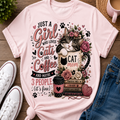 A light pink graphic t-shirt featuring a cute cat and coffee theme, styled in a flat-lay arrangement with white sneakers, blue jeans, a plant, and a straw hat on a wooden surface.