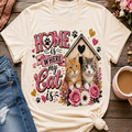 A cream-colored graphic t-shirt featuring a cute design of two cats, a birdhouse, and the text 'HOME is WHERE my Cat is' in pink and black lettering, surrounded by flowers and paw prints. The t-shirt is styled in a flat-lay arrangement on a wooden surface next to a cup of coffee and blue jeans.