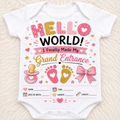 A white short-sleeve baby bodysuit laid flat on a soft beige textured background, printed with a colorful 'Hello World! I Finally Made My Grand Entrance' graphic featuring pink, gold, and black text, hearts, stars, pacifier, footprints, and a bow.