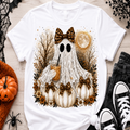 A white short-sleeve T-shirt laid flat on a light wood surface, featuring a large Halloween graphic of a ghost in a white lace dress holding an iced coffee, surrounded by pumpkins, autumn leaves, bare trees, a full moon, and leopard-print bows. The shirt is styled with seasonal props including a small pumpkin, black spider decoration, lace trim, folded dark jeans, and black sneakers.