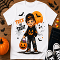 A white short-sleeve graphic T-shirt laid flat on a wooden surface with Halloween props. The shirt features a cartoon child in a black hoodie and cap holding a pumpkin bucket, with an orange full moon, white ghost, bats, spiderwebs, and the words “TRICK OR TREAT.