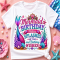 A white short-sleeve t-shirt with a colorful mermaid-themed birthday graphic, styled in a flat-lay on a pink background with seashells, starfish, and a mermaid tail.