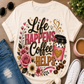 A cream-colored graphic t-shirt laid flat on a wooden surface, styled with a pair of blue jeans, white sneakers, sunglasses, and a pink coffee mug. The t-shirt features a colorful graphic with the text 'Life HAPPENS, Coffee HELPS' surrounded by pink and white roses, coffee beans, and a pink coffee cup with a gold heart.