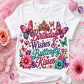 A white short-sleeve T-shirt laid flat on a pink floral surface, featuring a colorful decorative graphic with butterflies, flowers, hearts, and the text 'Birthday Wishes & Butterfly Kisses.' The shirt is styled with pink roses, glitter, and decorative props around it.