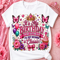 A white short-sleeve t-shirt with a colorful birthday graphic print, styled flat on a pink glittery background with decorative flowers and a tiara.
