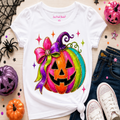A white short-sleeve T-shirt laid flat on a light wood surface, featuring a large colorful jack-o'-lantern graphic with a pink and purple bow. The shirt is styled with jeans, white sneakers, glittery pumpkins, spider decorations, and confetti.
