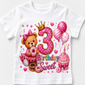 A white short-sleeve baby t-shirt featuring a colorful 3rd birthday graphic with a pink teddy bear, donuts, balloons, a cupcake, and a large pink number 3 with a gold crown.