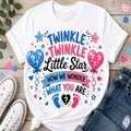 A white graphic T-shirt laid flat on a light wood surface, styled with white sneakers, blue jeans, greenery, and pastel confetti. The shirt features the phrase “TWINKLE TWINKLE Little Star HOW WE WONDER WHAT YOU ARE” in blue, pink, and black lettering, with balloons, stars, hearts, and baby footprints.