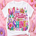 A white short-sleeve T-shirt laid flat on a pink background with decorative flowers and butterflies. The shirt features a colorful graphic reading 'Born to Flutter Born to SHINE' with a crown, butterflies, hearts, and flowers.