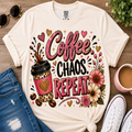 A cream-colored graphic t-shirt featuring the text 'Coffee CHAOS REPEAT.' in pink, black, and glittery lettering, styled in a flat-lay with a coffee cup, flowers, sunglasses, jeans, and a mug on a wooden surface.