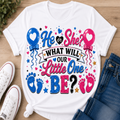 A white graphic T-shirt laid flat on a wood surface with white sneakers and blue jeans as styling props. The shirt features colorful text reading 'He or She? WHAT WILL OUR Little One BE?' with blue and pink balloons, hearts, stars, baby footprints, and glitter-style lettering.