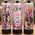 Three insulated tumbler cups with black speaker bases are shown on a wooden surface against a softly blurred indoor background. The tumblers feature colorful cat illustrations, flowers, butterflies, hearts, and decorative text including 'Life is Better with Cats' and 'Paw Stive Vibes.