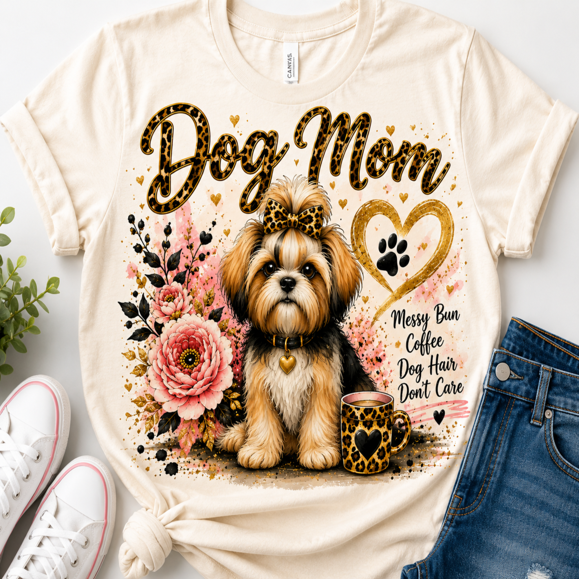 A cream-colored graphic t-shirt featuring a Shih Tzu dog, pink flowers, and the text 'Dog Mom' in leopard-print lettering. The shirt is styled in a flat-lay arrangement with white sneakers, a green plant, and blue denim jeans on a white background.