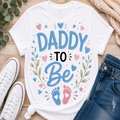 A white graphic T-shirt laid flat on a wooden surface with styled props including white sneakers, blue jeans, eucalyptus leaves, and a beige knit blanket. The shirt reads “DADDY TO Be” in blue and black lettering with pink and blue baby footprints and heart/star accents.