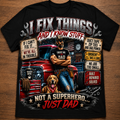 A black short-sleeve graphic t-shirt laid flat on a brown wooden surface. The shirt features a large, detailed graphic print with the text 'I FIX THINGS AND I KNOW STUFF' and 'NOT A SUPERHERO.