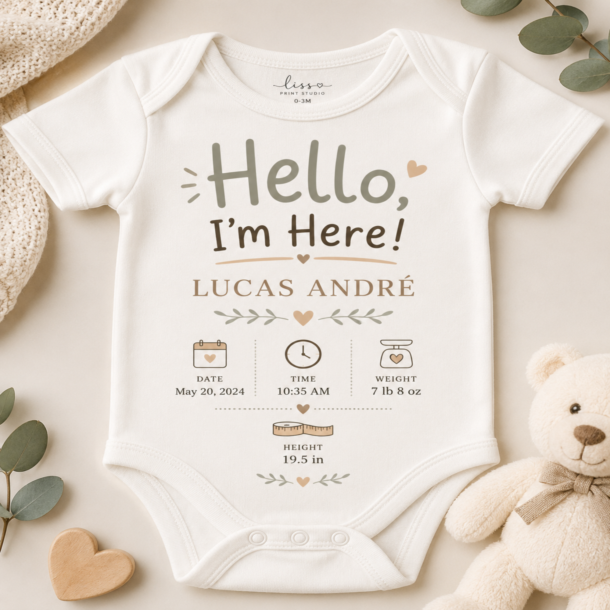 A white short-sleeve baby bodysuit laid flat on a light beige surface, printed with the phrase 'Hello, I'm Here!' and personalized baby details for Lucas André. The bodysuit is styled with a knit hat, wooden hearts, eucalyptus leaves, and a teddy bear.