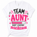 A white short-sleeve t-shirt with a graphic print that reads 'TEAM AUNT BABY LOADING CAN'T WAIT TO MEET YOU!' in black and pink lettering with heart and star accents, shown on a plain white background.
