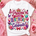 A white short-sleeve T-shirt with a colorful glitter-style birthday graphic reading 'Princess It's My Birthday Let's Celebrate!' surrounded by flowers, butterflies, bows, and balloons, styled on a pink furry surface with pink roses and a tiara.