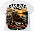 White short-sleeve crewneck t-shirt featuring a large, colorful graphic print on the front. The graphic depicts a man in an Adirondack chair fishing by a lake at sunset, with humorous text overlays like 'OFF DUTY Dad' and 'NOT RETIRED.