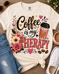 A cream-colored graphic t-shirt with a 'Coffee is my THERAPY' design, styled in a flat-lay arrangement with blue jeans, a white sneaker, sunglasses, and a coffee cup on a wooden background.