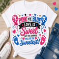 A white graphic t-shirt laid flat on a light wood surface, styled with white sneakers, blue jeans, and small pink and blue heart accents. The shirt features a colorful graphic with the text 'PINK or BLUE LOVE IS Sweet BUT THIS SURPRISE WILL BE THE Sweetest!' alongside pink and blue balloons, hearts, stars, and baby footprints.