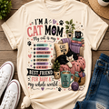 A cream-colored graphic t-shirt featuring a colorful 'Cat Mom' design, styled in a flat-lay arrangement with white sneakers, blue jeans, a straw hat, and plants on a wooden floor.