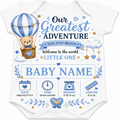 A white short-sleeve baby bodysuit featuring a blue and navy blue graphic print with a teddy bear in a hot air balloon, clouds, stars, and baby milestone icons. The text reads 'Our Greatest ADVENTURE HAS JUST BEGUN Welcome to the world LITTLE ONE BABY NAME' with placeholder date, time, weight, and length fields at the bottom.