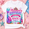 A white short-sleeve kids' t-shirt with a colorful mermaid birthday graphic, styled in a flat-lay on a pink textured background with seashells and starfish.