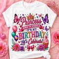 A white short-sleeve T-shirt laid flat on a pink glittery background with decorative flowers, a tiara, and balloons. The shirt features a large colorful birthday graphic reading 'Princess Born to Sparkle It's My Birthday Let's Celebrate!' with butterflies, flowers, a cupcake, and decorative text.