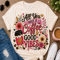 A cream-colored graphic t-shirt laid flat on a wooden surface, surrounded by lifestyle props including blue jeans, white sneakers, sunglasses, a cup of coffee, and a small plant. The t-shirt features a colorful graphic with the text 'All You NEED IS Coffee & GOOD VIBES' and an illustration of a coffee cup, flowers, and hearts.