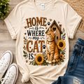 A cream-colored graphic t-shirt featuring an illustration of an orange tabby cat, sunflowers, a wooden house, and paw prints with the text 'HOME IS WHERE MY CAT IS'. The t-shirt is styled in a flat-lay arrangement on a wooden surface alongside white sneakers, blue jeans, and a small plant.