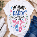 A white graphic t-shirt with the text 'MOMMY AND DADDY Can't Wait TO Meet YOU!' in black, blue, and pink lettering, surrounded by baby footprints, hearts, and floral details. The shirt is styled in a flat lay on a wooden surface with white sneakers, blue jeans, and a knit blanket.