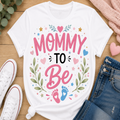 A white graphic t-shirt laid flat on a wooden surface, styled with white sneakers, blue jeans, a pink knit blanket, and greenery. The t-shirt features a colorful graphic that reads 'MOMMY TO Be' with pink and blue baby footprints and floral accents.