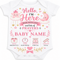 A white short-sleeve baby bodysuit with a pink and gold 'Hello, I'm Here Made with Love and lots of PRAYERS BABY NAME' graphic on the front, featuring cute cartoon stars, a crescent moon, and baby milestone icons at the bottom.
