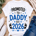 A white graphic T-shirt laid flat on a wooden surface with white sneakers, blue jeans, and a knit sweater around it. The shirt reads “PROMOTED TO DADDY EST.