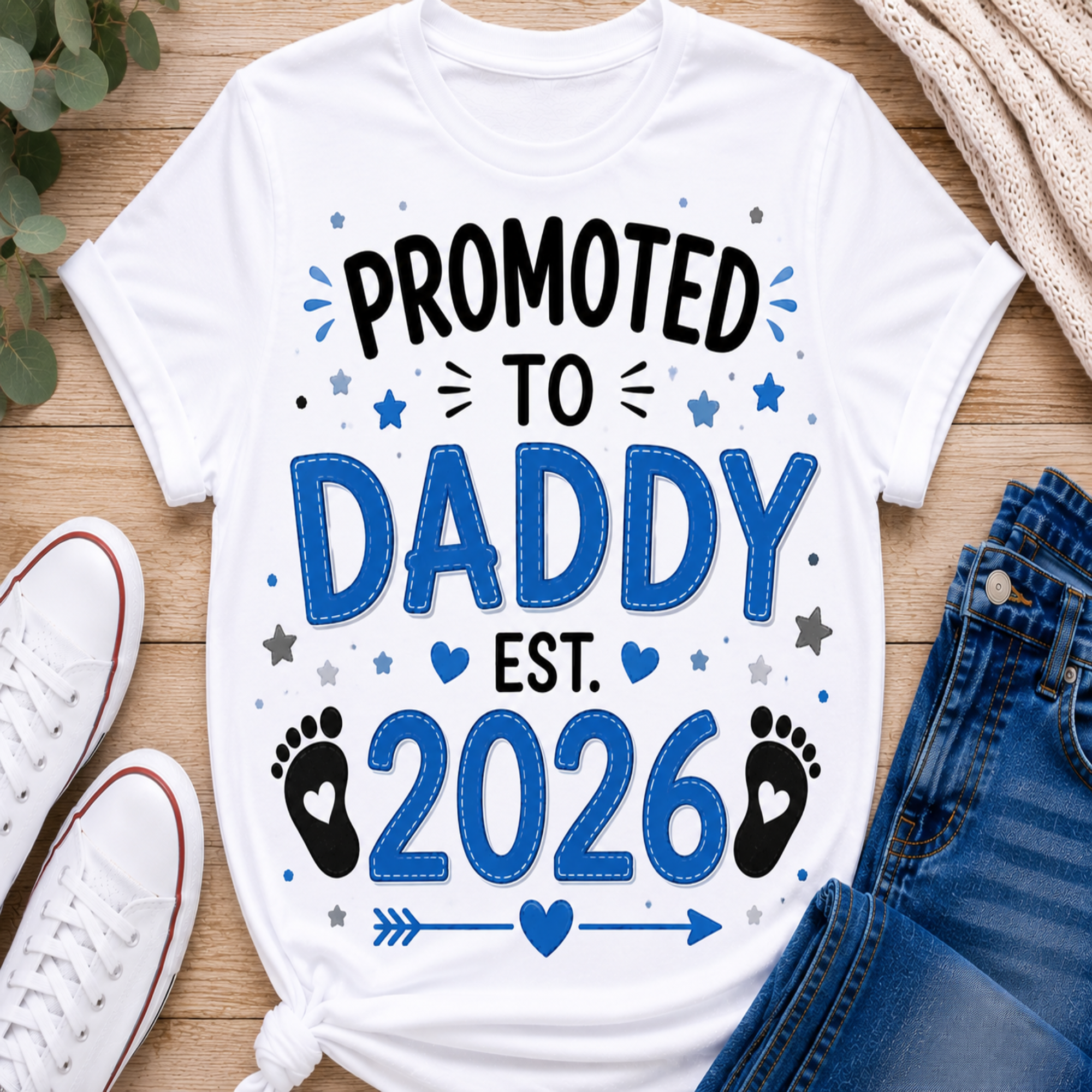 A white graphic T-shirt laid flat on a wooden surface with white sneakers, blue jeans, and a knit sweater around it. The shirt reads “PROMOTED TO DADDY EST.
