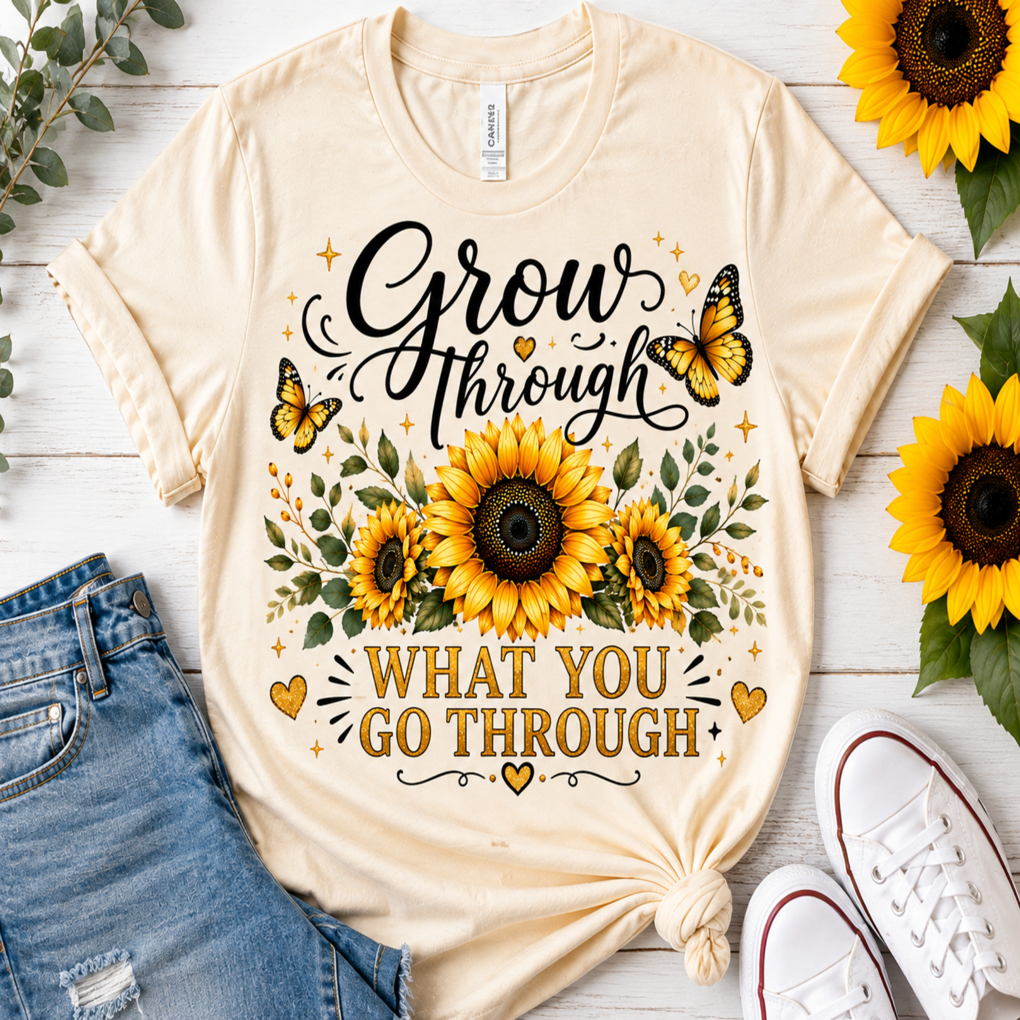 A cream-colored graphic t-shirt with a sunflower and butterfly design that reads 'Grow through what you go through'. The shirt is styled in a flat-lay arrangement on a white wood background, accompanied by blue denim shorts, white sneakers, and sunflowers.