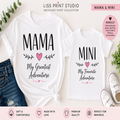 Matching white t-shirts for Mama and Mini from Liss Print Studio, featuring black text and pink heart accents. The larger shirt reads 'MAMA My Greatest Adventure' and the smaller matching shirt reads 'MINI My Favorite Adventure'.