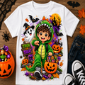 A white short-sleeve T-shirt laid flat on a wooden surface, featuring a colorful Halloween graphic of a child in a green dinosaur costume surrounded by pumpkins, bats, a ghost, and a 'Trick or Treat' sign. The shirt is styled with jeans, black sneakers, a pumpkin bucket of candy, and autumn leaves.