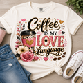 A white graphic T-shirt laid flat on a light wood surface, styled with blue jeans, a pink cup, and white sneakers. The shirt features a large decorative coffee-themed print reading 'Coffee is my love language' with pink hearts, flowers, a coffee cup, and gold glitter-style accents.
