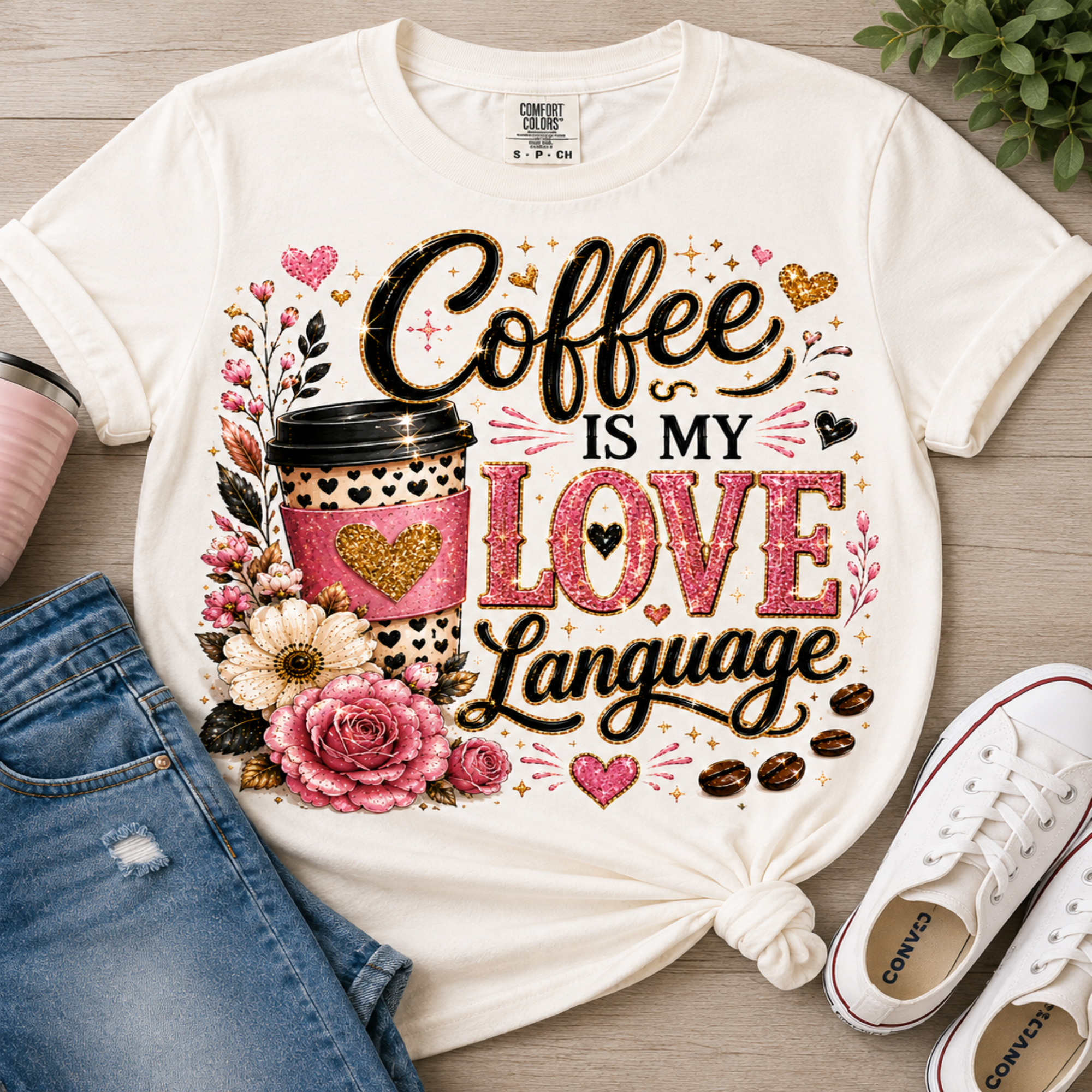 A white graphic T-shirt laid flat on a light wood surface, styled with blue jeans, a pink cup, and white sneakers. The shirt features a large decorative coffee-themed print reading 'Coffee is my love language' with pink hearts, flowers, a coffee cup, and gold glitter-style accents.