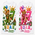 Two white short-sleeve T-shirts shown side by side, each printed with a colorful cartoon birthday design featuring a teddy bear riding a dinosaur, a large number 7 with a crown, balloons, flowers, and the text 'ROAR I'm Seven'.