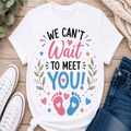 A white graphic t-shirt laid flat on a wooden surface, surrounded by white sneakers, blue jeans, and a knitted sweater. The t-shirt features a colorful graphic that reads 'WE CAN'T Wait TO MEET YOU!' with baby footprints and hearts.
