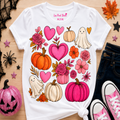 A white graphic t-shirt with a colorful Halloween print featuring pumpkins, hearts, flowers, and ghosts, styled in a flat-lay with jeans, pink sneakers, and autumn leaves on a wooden surface.