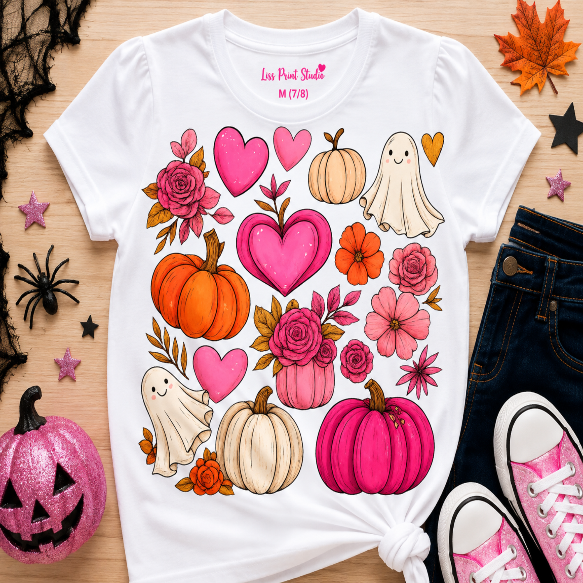 A white graphic t-shirt with a colorful Halloween print featuring pumpkins, hearts, flowers, and ghosts, styled in a flat-lay with jeans, pink sneakers, and autumn leaves on a wooden surface.