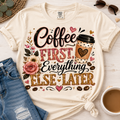 A cream-colored graphic t-shirt with a 'Coffee First, Everything Else Later' design, styled in a flat-lay arrangement with blue jeans, sunglasses, a cup of coffee, and a woven mat on a light wood background.